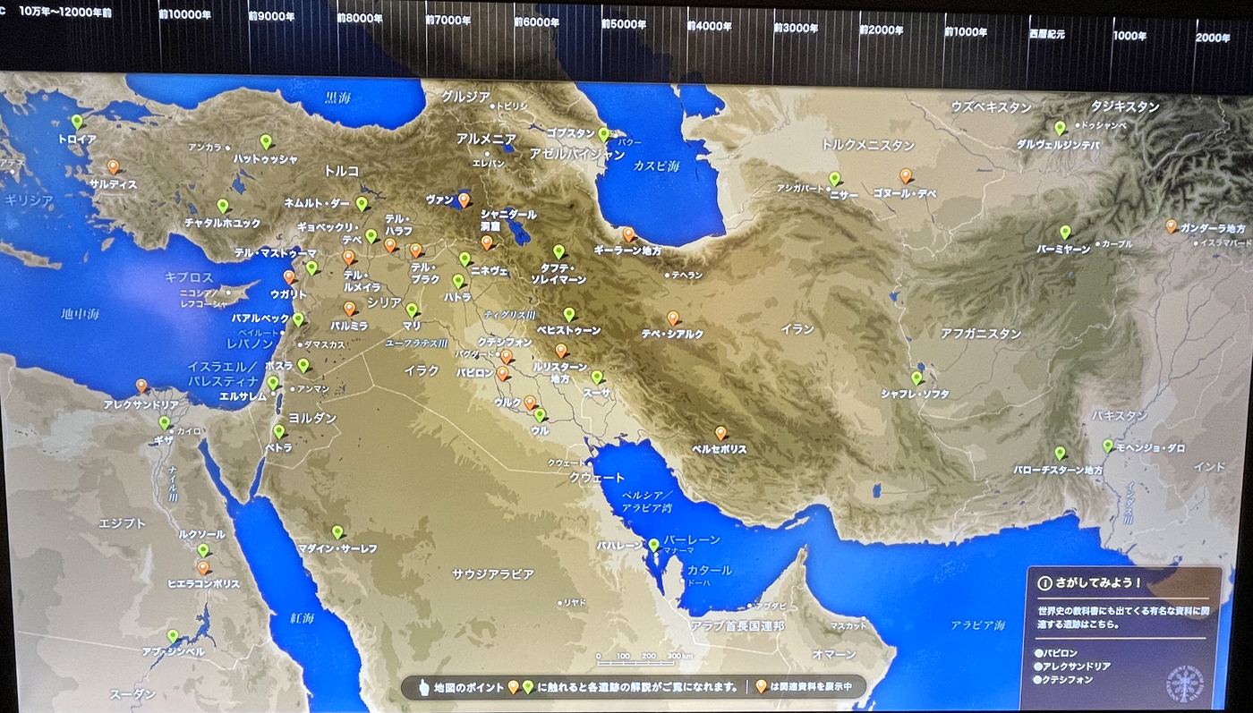 博物館の大型画面に映る古代オリエントの地図。上端に年表の帯があり、地図の上に遺跡を示すピンが並んでいる。