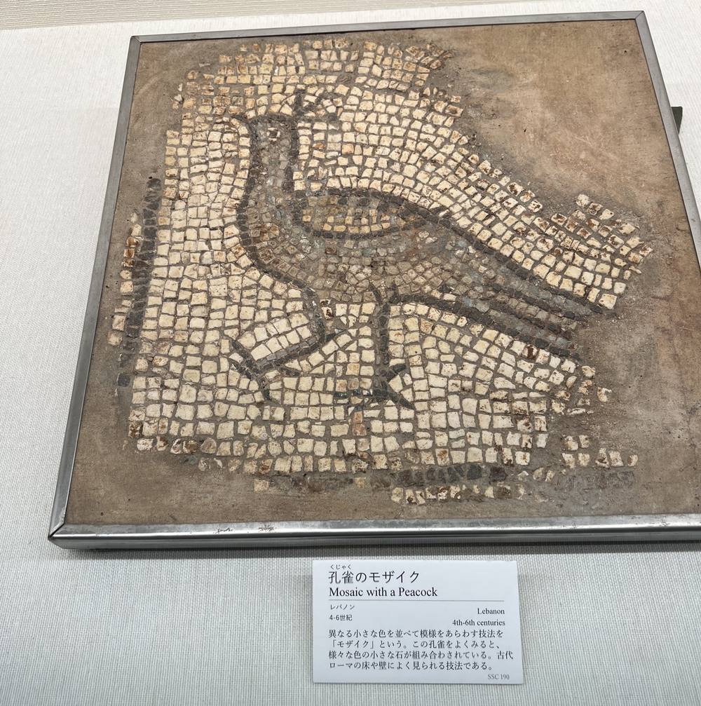 小さな石片を並べて描かれた孔雀のモザイク。金属の枠に収められ、下に展示の解説札が置かれている。